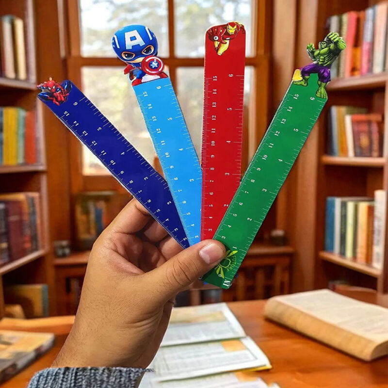 A hand holding up four Superhero Scale Rulers with different Marvel Avengers characters on top, including Captain America, Iron Man, Spider-Man, and Hulk, set against a blurred background of bookshelves.