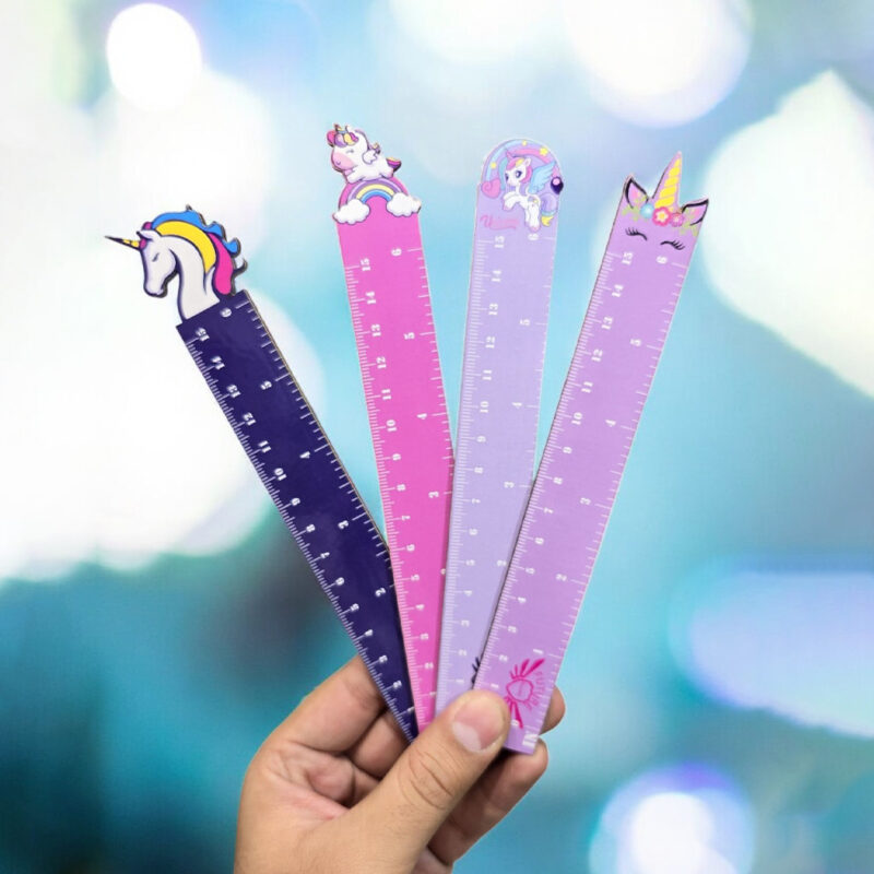 A hand holding up four colorful Unicorn Scale Rulers with different unicorn designs on top, against a blurred, bright background.