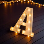 A white 24cm LED Marquee Letter Light in the shape of the letter 'A' illuminated with warm white lights, sitting on a dark wooden surface.