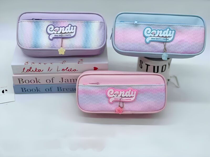 Three pastel-colored Candy Pouches (lavender, blue, pink) with iridescent fronts and lollipop zipper charms, stacked on white books, on a bright surface.
