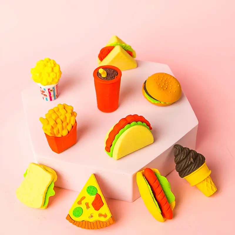 A collection of colorful Food Eraser Pack items, including miniature french fries, hamburgers, tacos, pizza, ice cream, and drinks, arranged on pink and white hexagonal platforms.