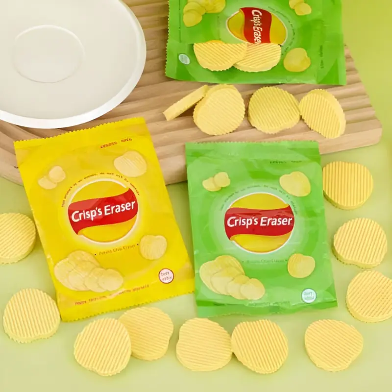 Several yellow and green mini "Crisp's Eraser" chip bags, with numerous yellow crinkle-cut Kawaii Chips Erasers scattered around them on a green and white surface.