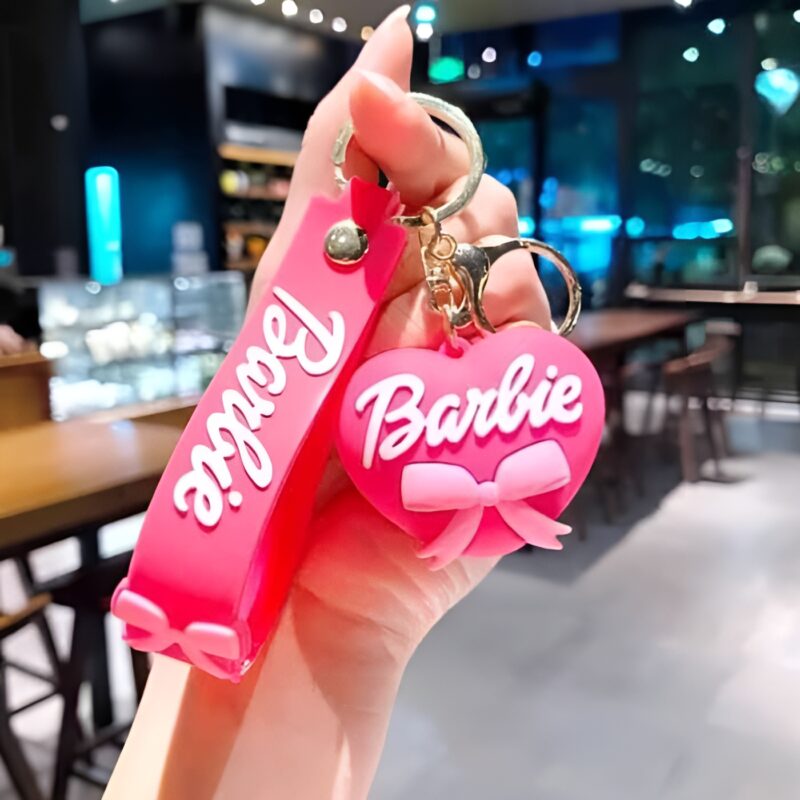 A hand holding a vibrant pink Barbie Heart Keychain, consisting of a heart-shaped charm and a strap, both featuring the Barbie logo and a bow design.